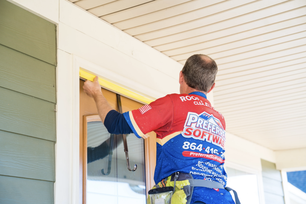 A person in a colorful Preferred SoftWash shirt is applying yellow tape above a door frame on a green house. They have a tool belt around their waist and are standing on a step ladder under a white ceiling.